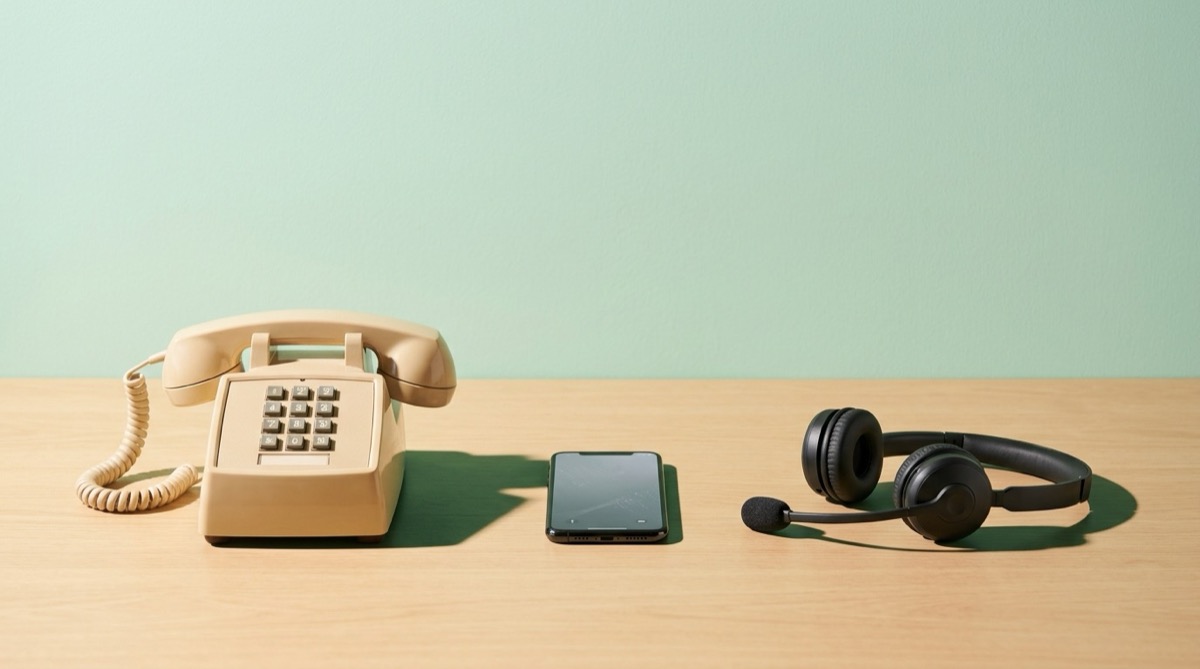 Three telephones of different kinds side by side on a calm table, a desk phone, a mobile and a headset